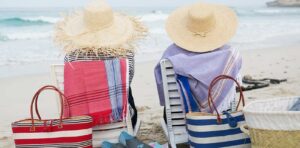Two people in sun hats sitting on beach chairs with striped towels and bags by the ocean.