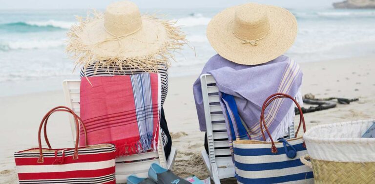 Two people in sun hats sitting on beach chairs with striped towels and bags by the ocean.