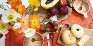 Colorful table setting with flowers, fruits, bagels, and wooden bowls on an orange cloth.